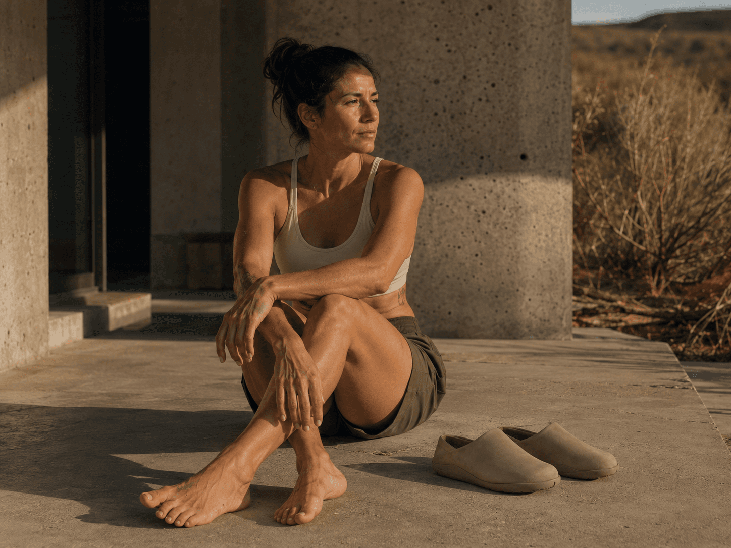 Athlete seated barefoot on a concrete pad in desert light, sand mules beside her.