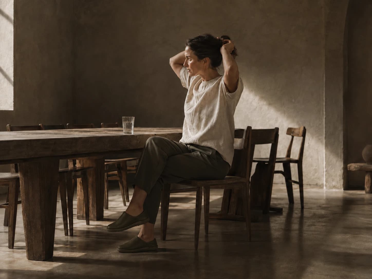 Hospitality worker sitting at a wood table at the end of a shift, taking her hair back, wearing soft leather slip-ons.
